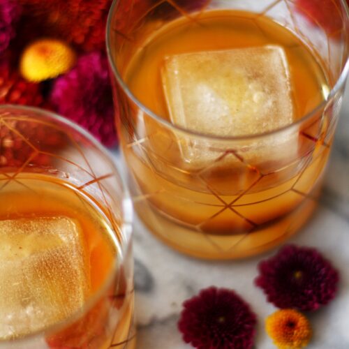Two glasses of amber-colored drinks with large square ice cubes sit on a marble surface, surrounded by vibrant red and purple flowers.