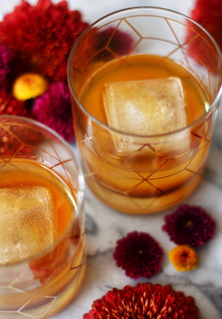 Two glasses of amber-colored drinks with large square ice cubes sit on a marble surface, surrounded by vibrant red and purple flowers.