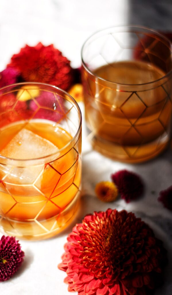 Two glasses of orange-colored drink with ice, featuring geometric patterns, placed on a white surface surrounded by bright red and magenta flowers. Bright sunlight highlights part of the scene.