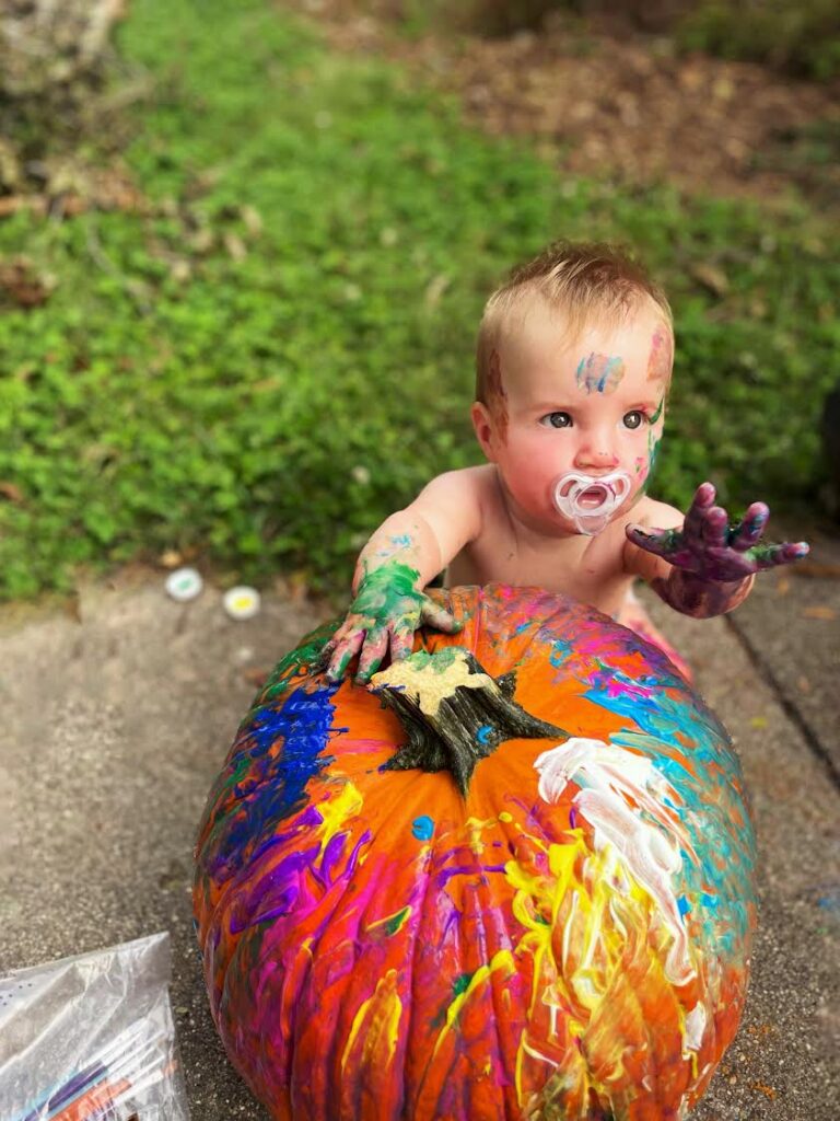 A baby with a pacifier, covered in colorful paint, reaches out while sitting behind a large pumpkin that is also painted with bright swirls. The scene is outdoors on a sidewalk next to some grass.