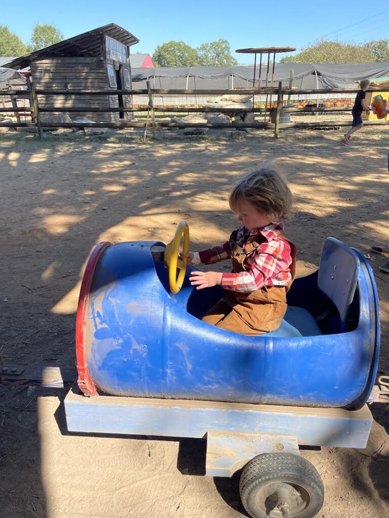 A young child in a plaid shirt and overalls sits in a small blue barrel-shaped cart with a yellow steering wheel at an outdoor farm or ranch. A wooden building and fences are visible in the background.