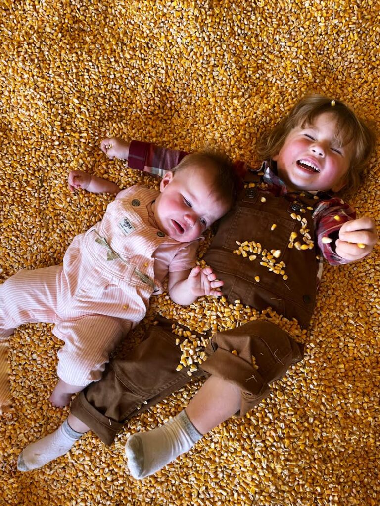 Two young children lie on a large pile of corn kernels. The older child is smiling and playing with the corn, while the younger baby looks up with a curious expression. Both are dressed in overalls.