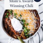 A bowl of chili topped with shredded cheese, green onions, and a dollop of sour cream sits on a striped towel, with a spoon and a label reading Mom’s Award-Winning Chili: try this award winning chili recipe for a hearty dinner.
