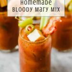A glass of homemade Bloody Mary, made with zesty homemade bloody mary mix and garnished with celery, olives, cheese, and bacon, sits on a table. The text on the image reads “Easy Recipe: Homemade Bloody Mary Mix.”.