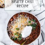 Overhead view of a bowl of award winning chili recipe topped with shredded cheese, sour cream, and chopped green onions, with a spoon in the bowl. Text overlay reads, “Mom’s Award-Winning Hearty Chili Recipe.”.