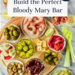 A round wooden platter with bowls of Bloody Mary garnishes—shrimp, olives, pickles, cheese, celery, and peppers—sits on marble. Text reads: Build Your Own Bloody Mary Bar for the ultimate customizable cocktail experience.