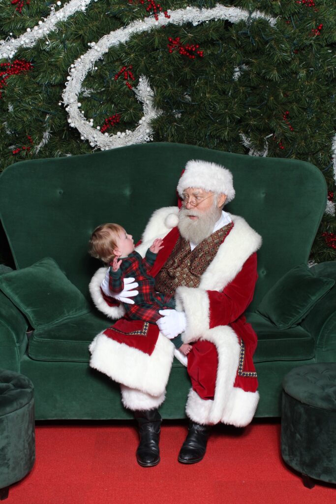 A man dressed as Santa Claus sits on a green velvet couch holding a young child in plaid pajamas. The background features festive holiday greenery and decorations.