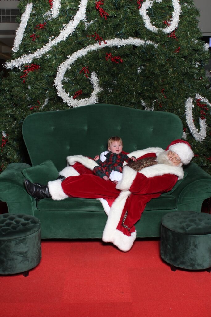 A toddler sits next to a reclining Santa Claus on a large green couch in front of a decorated Christmas tree, with red carpet and two green ottomans in the foreground.