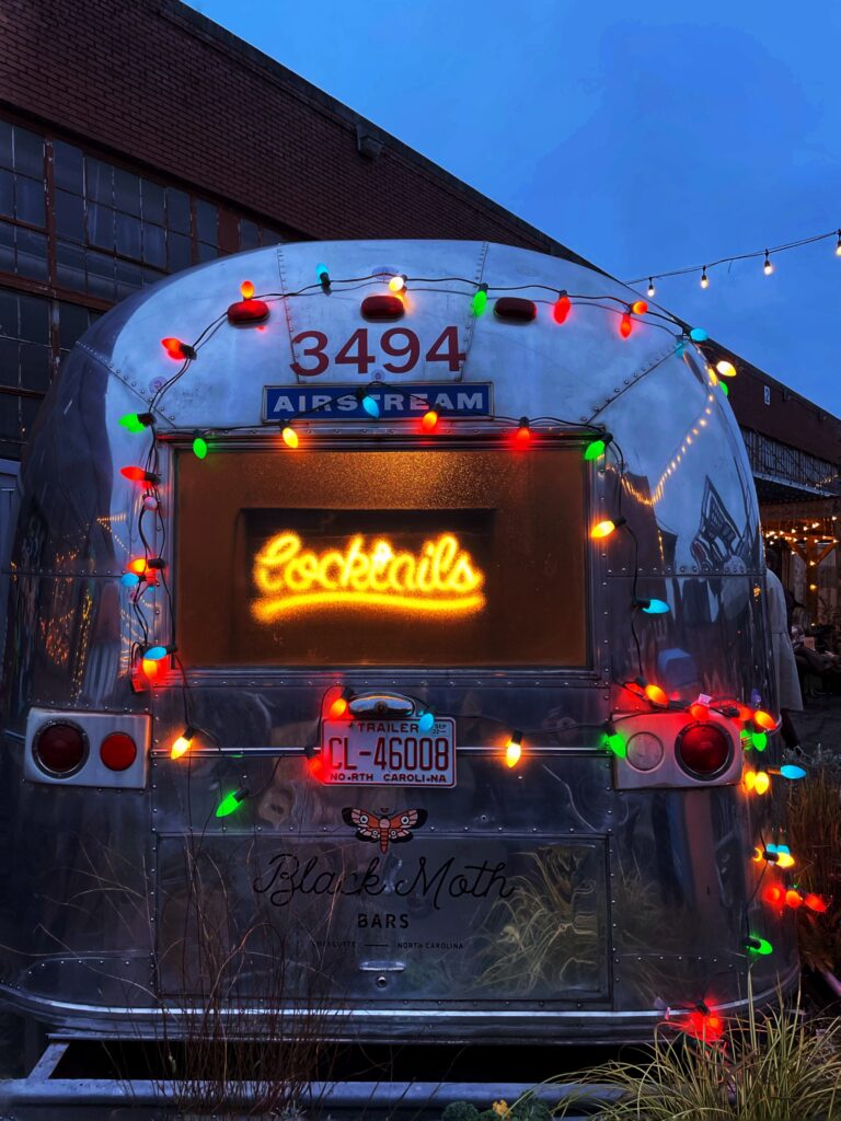 Airstream trailer decorated with colorful string lights and a glowing neon Cocktails sign in the window, parked near a brick building at dusk.