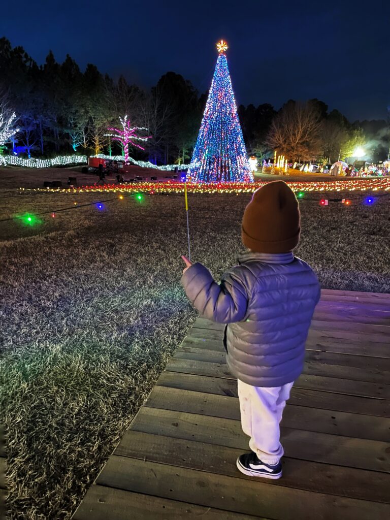 A small child in a brown hat and gray jacket stands on a wooden path at night, holding a yellow glow stick, gazing at a large, colorful, lit Christmas tree and festive lights in the distance.