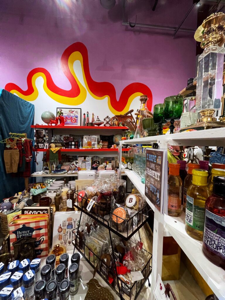 A colorful vintage store interior with shelves full of eclectic items like glassware, bottles, board games, and trinkets. A wavy red and yellow mural decorates the purple wall in the background.