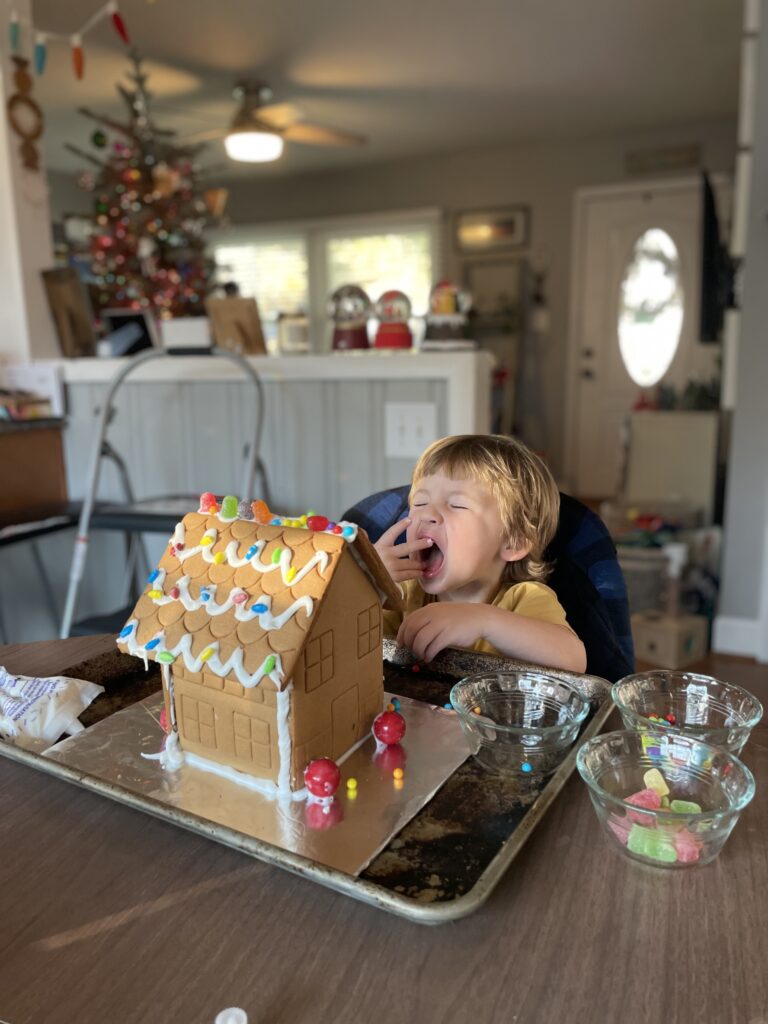 A young child sits at a table in front of a decorated gingerbread house, yawning or shouting with eyes closed. The table has bowls of candy, and a Christmas tree is visible in the background.