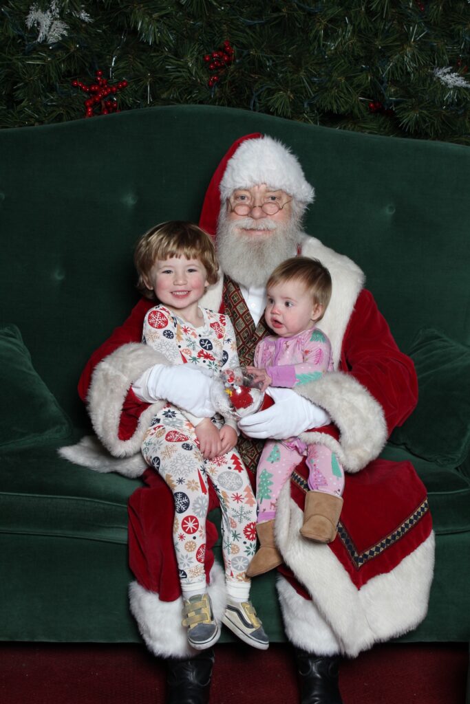 Santa Claus sits on a green couch holding two young children on his lap. One child smiles, wearing festive pajamas, while the other looks uncertain, also in pajamas. Greenery with red berries hangs in the background.