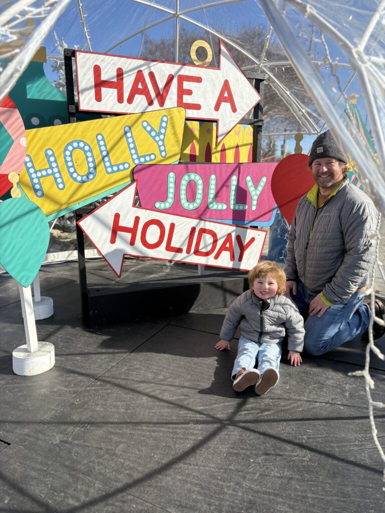 A man and a young child sit smiling in front of a colorful sign that reads “Have a Holly Jolly Holiday.” They are bundled in winter clothes, enjoying a festive outdoor setting with bright decorations.