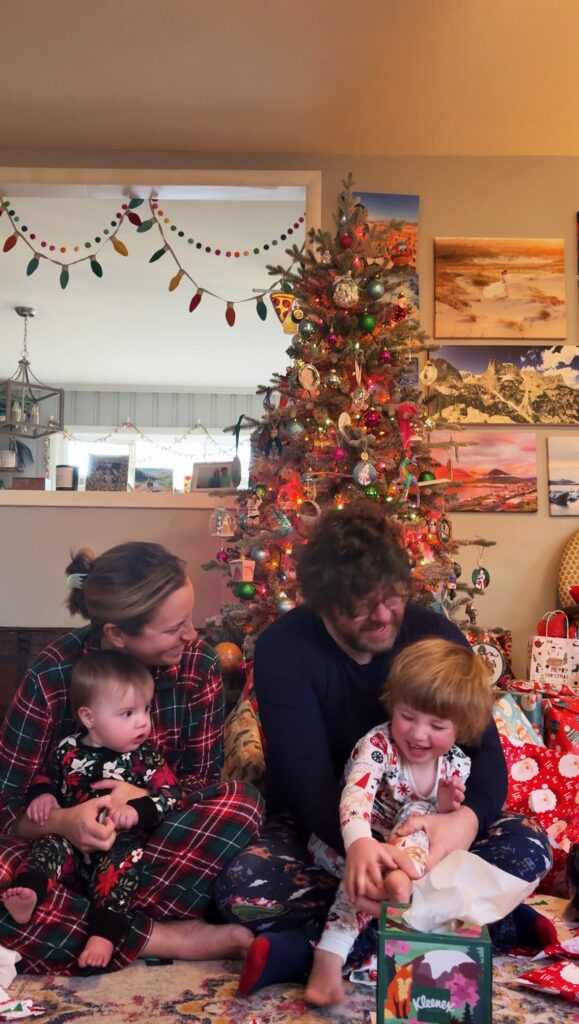 A family sits together in front of a decorated Christmas tree, surrounded by wrapped presents. Two adults smile at their two young children, all dressed in festive pajamas. The room is decorated for the holidays.
