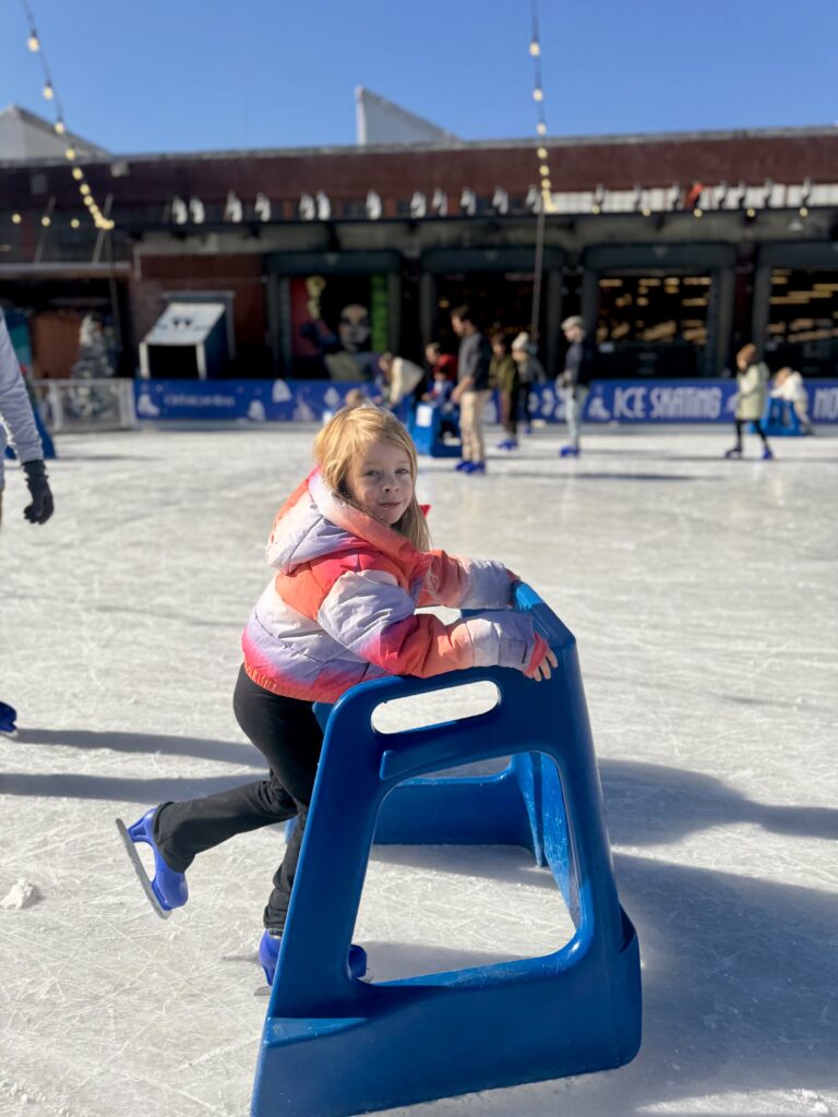 A young child in a pink and orange jacket uses a blue skating aid while ice skating outdoors on a sunny day. Other skaters and people are visible in the background.