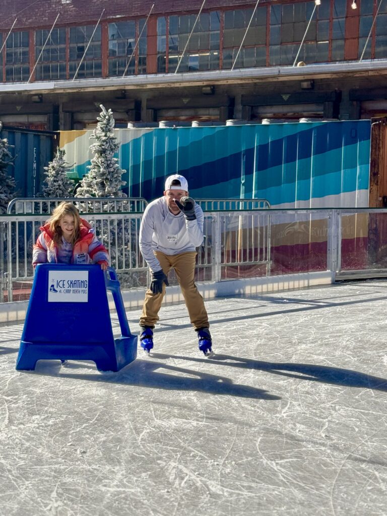 Two people ice skating outdoors on a sunny day; a child in a red jacket uses a blue ice skating aid, while an adult in a white hoodie skates beside her, both appearing to enjoy themselves.