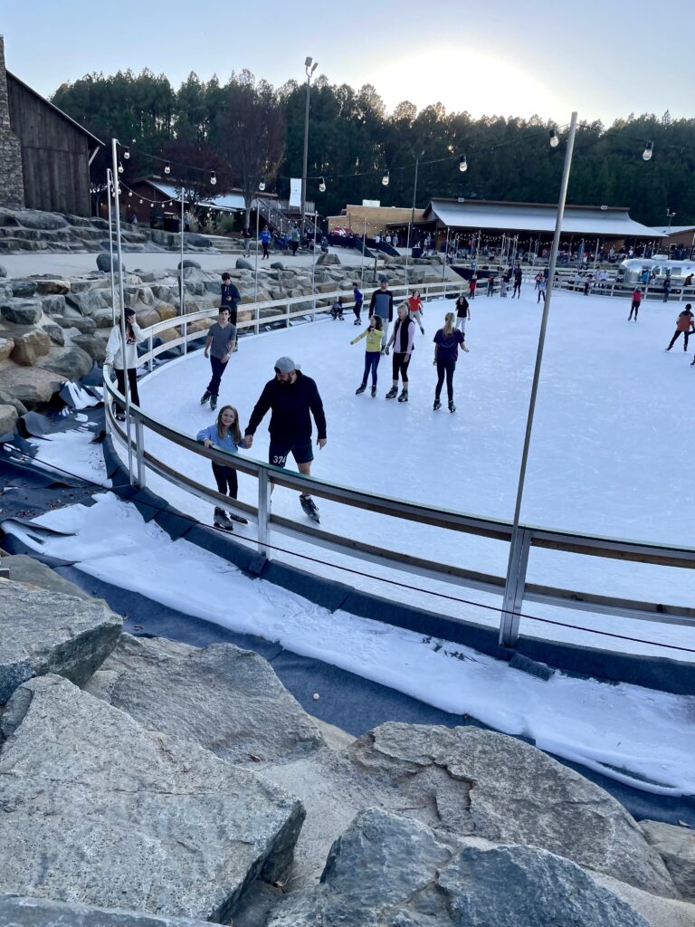People of various ages ice skating on an outdoor rink surrounded by rocks and wooden buildings, with trees in the background and a crowd watching from the sidelines.