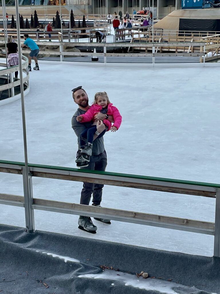 A man in ice skates smiles while holding a young girl, also in skates and a bright pink jacket, on an outdoor ice skating rink. They both appear happy and are standing near a wooden railing.