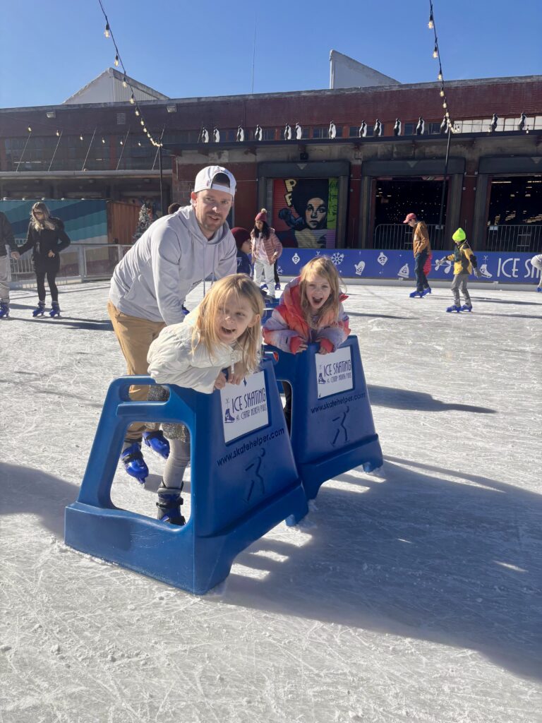 A man and two young girls are ice skating outdoors on a sunny day, using blue skating aids. Other skaters and a decorated building are visible in the background. Everyone looks happy and is enjoying the activity.