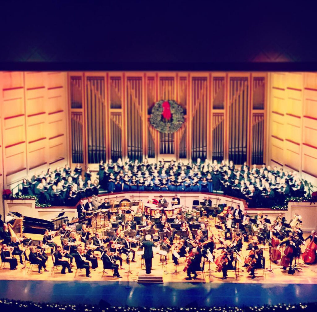 A symphony orchestra performs on stage with a large choir behind them. The stage is decorated with a festive wreath and warm lighting, creating a celebratory atmosphere in a concert hall.
