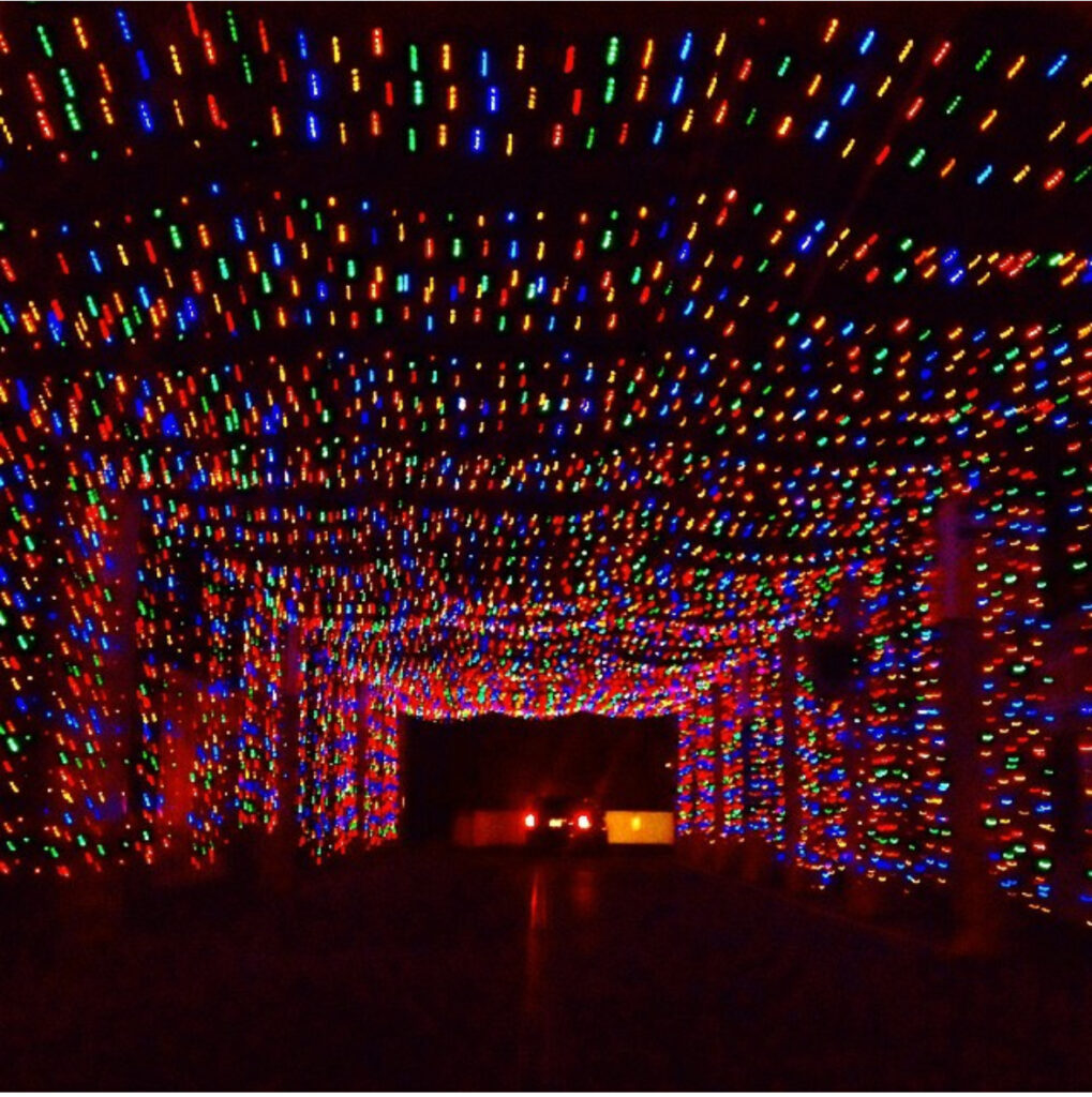 A tunnel is decorated with thousands of small, colorful holiday lights in red, blue, and green, creating a vibrant, festive canopy. Two cars with red taillights are visible driving through the tunnel.