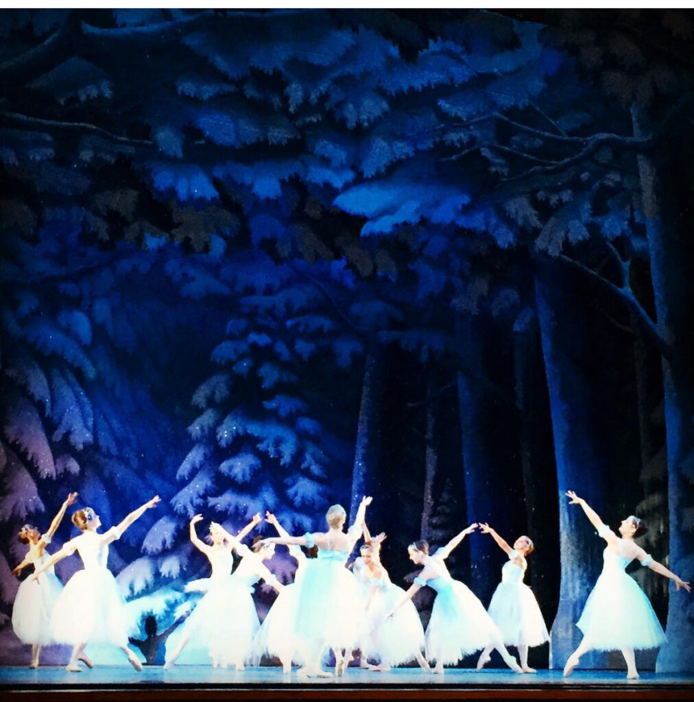 Ballet dancers in white tutus perform on stage, arms gracefully extended, against a backdrop of a dark, snowy forest with tall trees and blue-toned lighting, creating a magical winter scene.