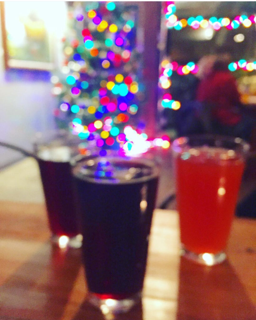 Three glasses of drinks on a wooden table in the foreground, with colorful holiday lights and a decorated Christmas tree blurred in the background, creating a festive atmosphere.
