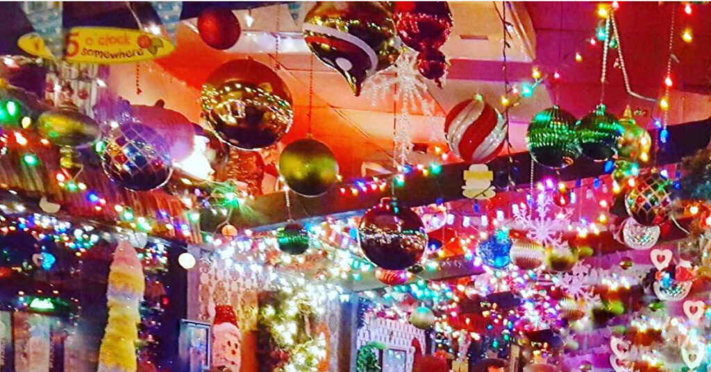 Colorful Christmas decorations fill a ceiling, featuring hanging ornaments, shiny baubles, snowflakes, and multicolored string lights, creating a festive and vibrant holiday atmosphere indoors.