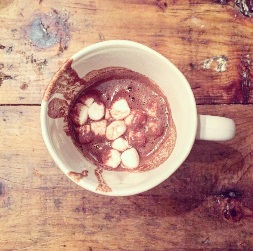 A white mug filled with hot chocolate topped with mini marshmallows sits on a rustic wooden table. Some hot chocolate has spilled onto the inside rim of the mug.