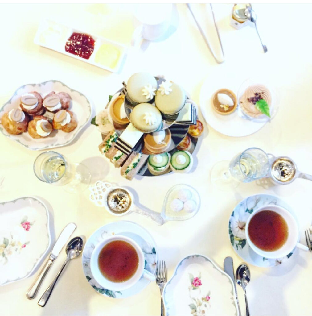 A bright, top-down view of an elegant afternoon tea setting with floral china, cups filled with tea, assorted pastries, finger sandwiches, macarons, scones, and small desserts on a white tablecloth.