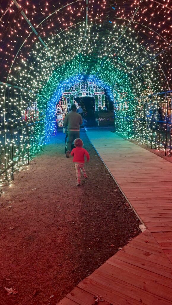 A small child in red runs toward an adult pushing a stroller under a tunnel of colorful holiday lights at night, with green, blue, and white lights creating a festive, glowing archway.