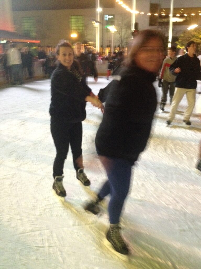 Two people ice skating, holding hands. One is facing the camera and smiling, while the other is turned away, appearing slightly blurred. There are other skaters and city lights in the background.