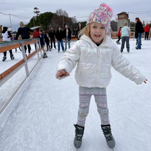 A young girl wearing a colorful knit hat, white puffy jacket, and patterned leggings enjoys ice skating in Charlotte, NC. She smiles with arms outstretched as people skate in the background under a cloudy sky.