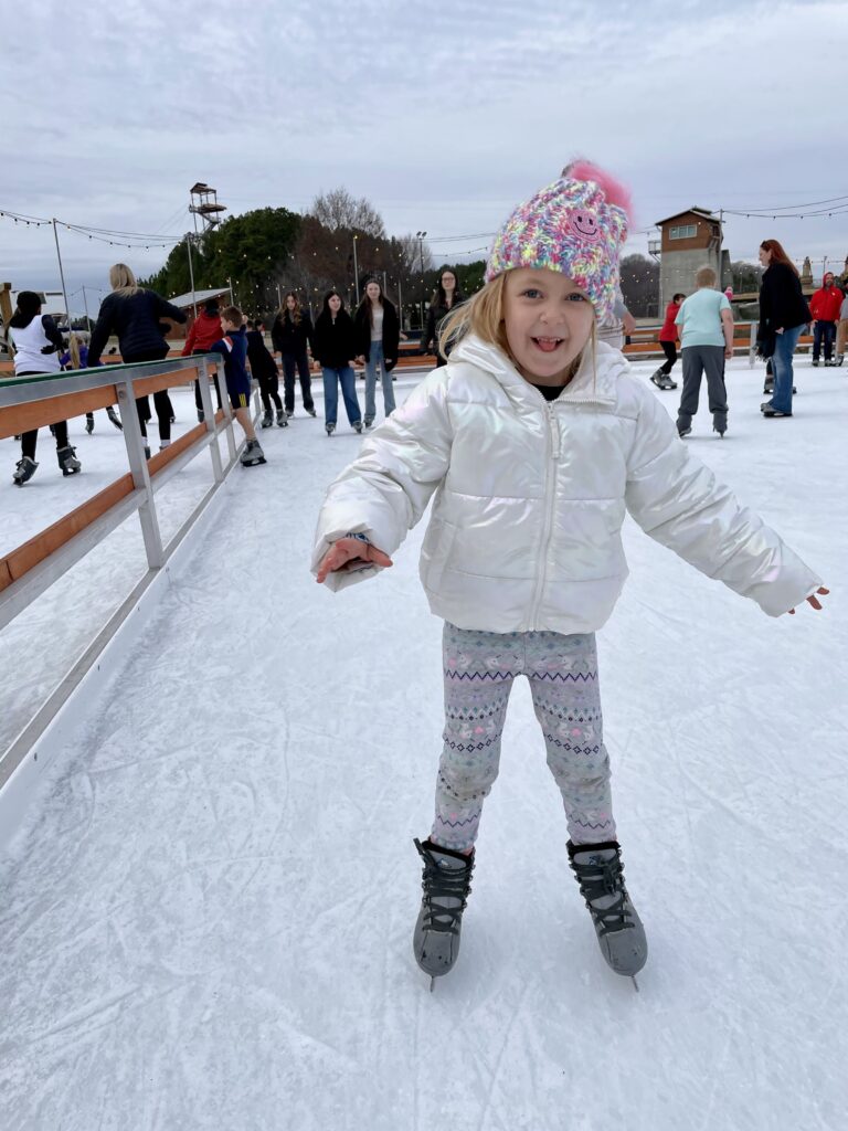 A young girl in a white jacket, patterned leggings, and a colorful hat smiles while ice skating outdoors. Other people are skating in the background under a cloudy sky.