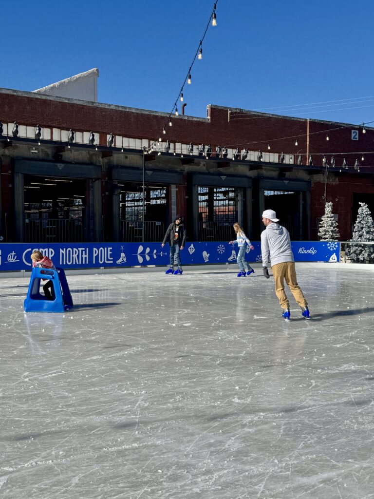 Four people ice skating at an outdoor rink on a sunny day. One child uses a blue skating aid, while three others skate nearby. Festive decorations and string lights are visible, with a brick building in the background.