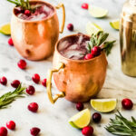 Two copper mugs filled with a dark red drink, garnished with cranberries and rosemary, sit on a marble surface with scattered cranberries, lime wedges, rosemary sprigs, and a gold cocktail shaker nearby.