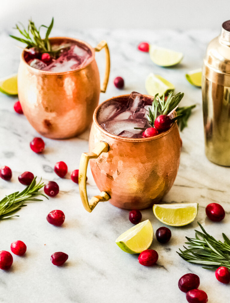 Two copper mugs filled with a dark red drink, garnished with cranberries and rosemary, sit on a marble surface with scattered cranberries, lime wedges, rosemary sprigs, and a gold cocktail shaker nearby.