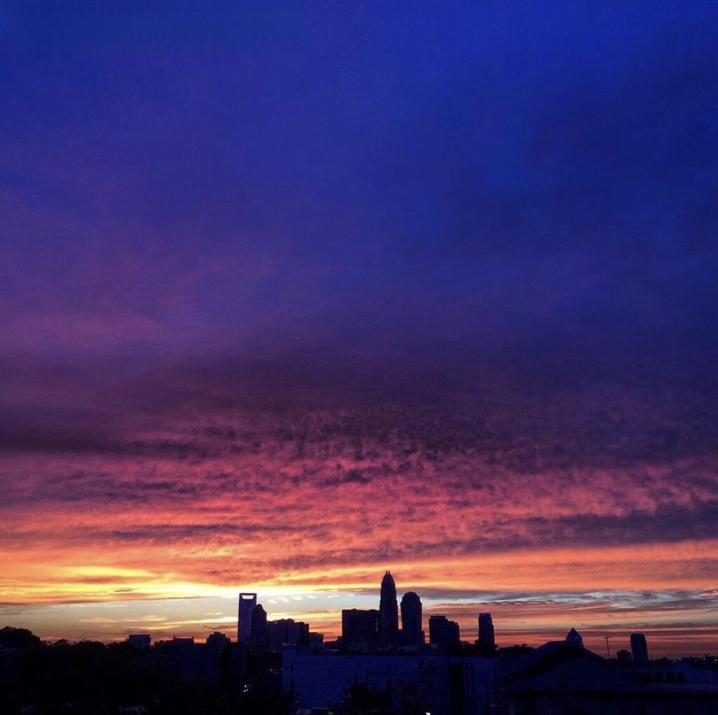 A city skyline is silhouetted against a dramatic sunset sky, with vibrant shades of orange, pink, purple, and deep blue blending above the buildings—a perfect scene to end a day spent exploring things to do in Charlotte.