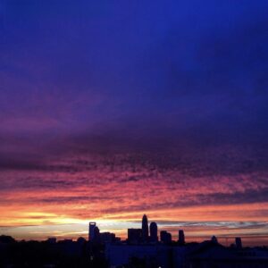 A city skyline is silhouetted against a dramatic sunset sky, with vibrant shades of orange, pink, purple, and deep blue blending above the buildings—a perfect scene to end a day spent exploring things to do in Charlotte.