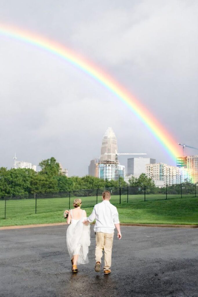 A couple walks hand in hand toward a city skyline under a bright rainbow, exploring things to do in Charlotte, with green grass and trees in the foreground and a tall building in the background.