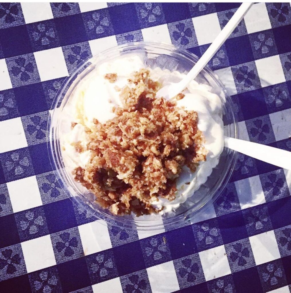 A clear plastic bowl of vanilla ice cream topped with a crumbly brown topping, with two white plastic spoons, sits on a blue and white checkered tablecloth—perfect for enjoying while exploring things to do in Charlotte.