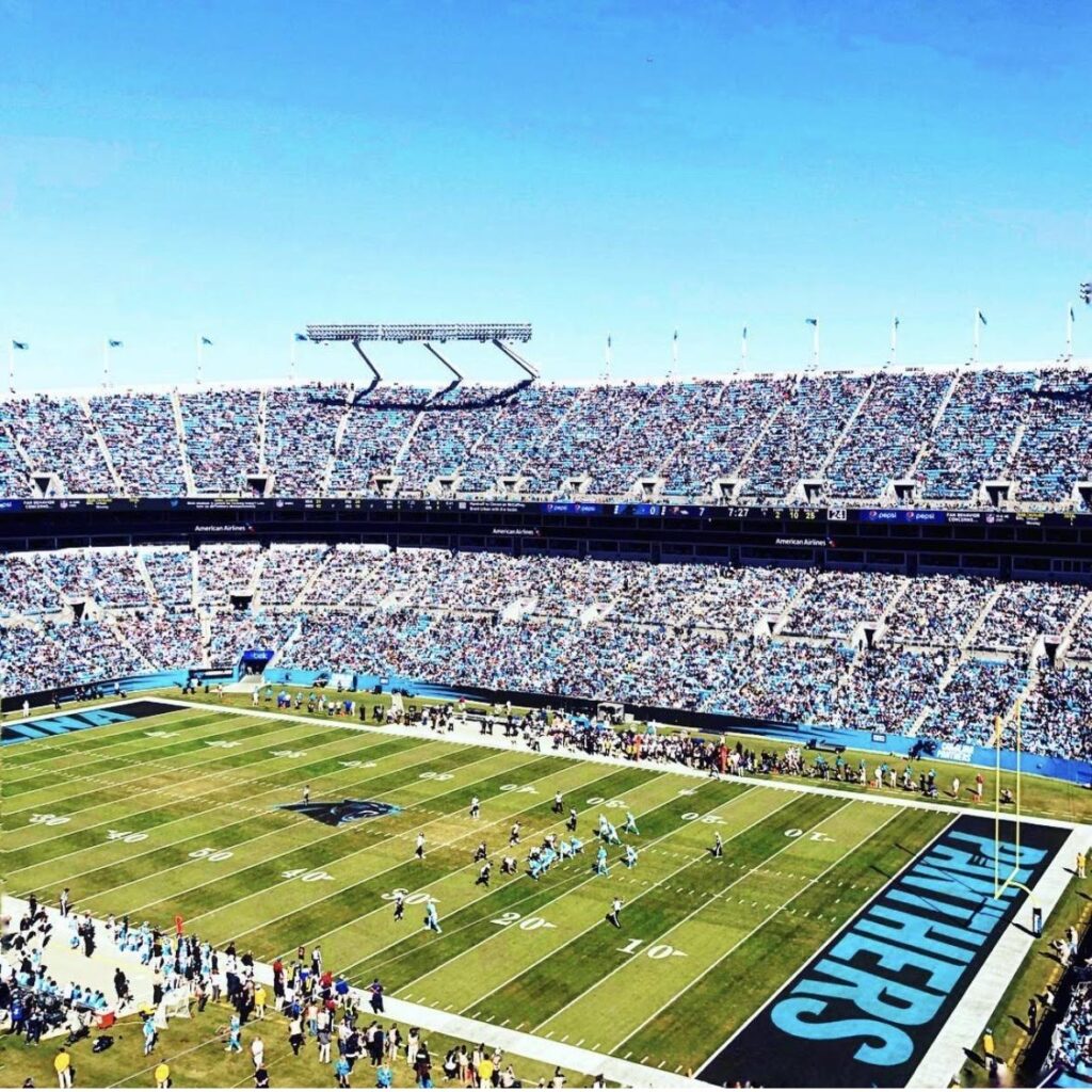 A large football stadium filled with spectators watching a game in progress—one of the top things to do in Charlotte. The end zone displays PANTHERS in bold letters as players are spread across the bright green field under a clear, blue sky.