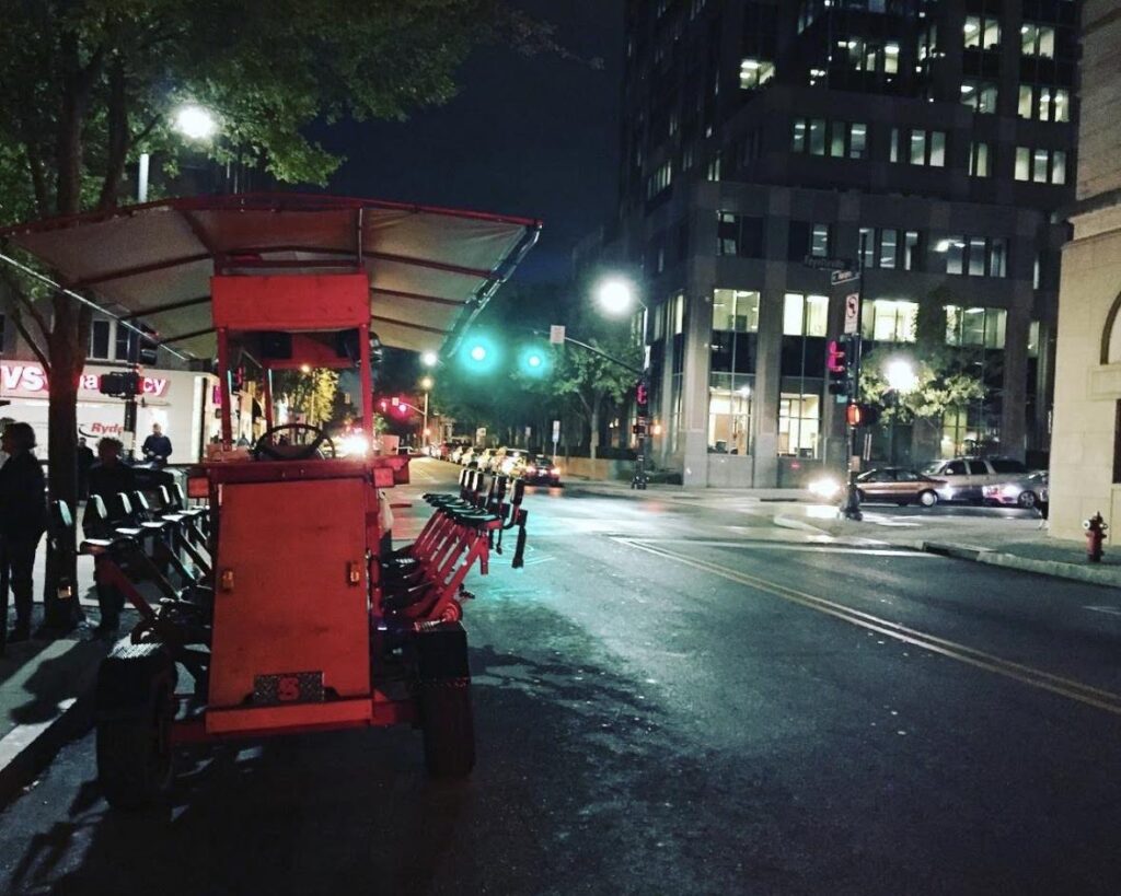 A pedal-powered party bike is parked on a city street at night, surrounded by buildings, streetlights, and traffic signals—one of the fun things to do in Charlotte. Some people stand nearby on the sidewalk, ready for the ride.