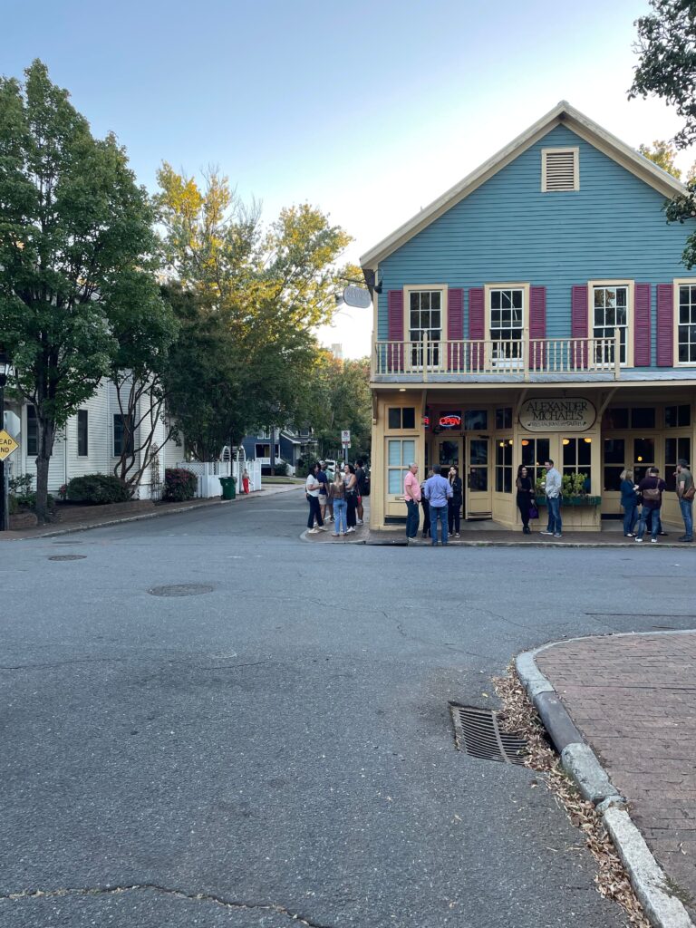 A group of people stands outside a blue and yellow two-story building with red shutters on a quiet, tree-lined street corner—a charming spot that could be among the hidden things to do in Charlotte's small towns.