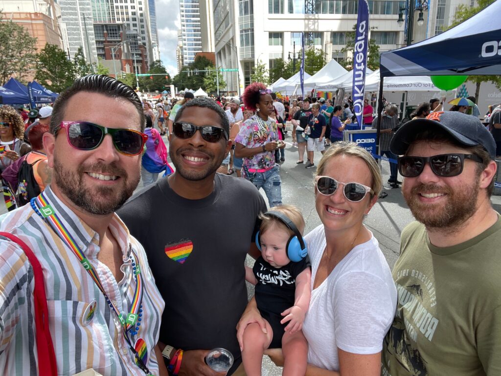 Five adults and a baby wearing headphones smile for a group selfie at an outdoor festival in Charlotte, surrounded by a crowd and colorful tents—one of the many fun things to do in Charlotte with vibrant city buildings in the background.