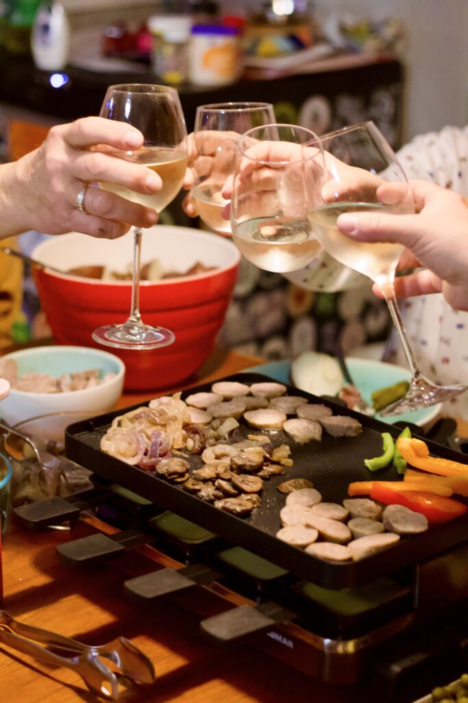 Four people raise glasses of white wine in a toast over a hot grill at a vibrant raclette dinner party, with sliced vegetables, meat, and colorful bowls of food surrounding the table.