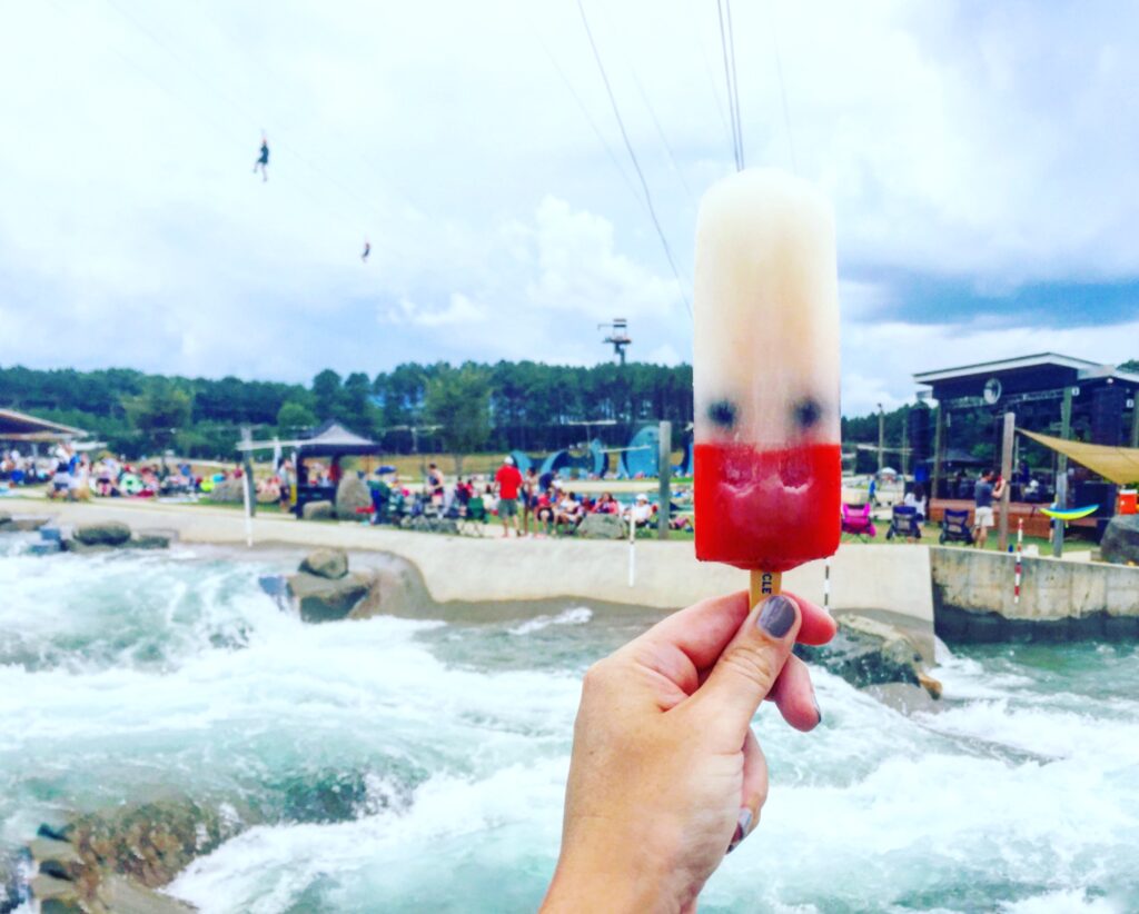 A hand holds a red, white, and blue popsicle in front of a whitewater rafting course—one of the top things to do in Charlotte—with people, buildings, and trees in the background under a cloudy sky.