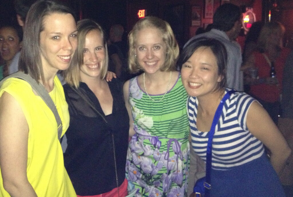 Four women smiling and posing together in a dimly lit indoor setting, possibly at a party or social event—one of the many fun things to do in Charlotte, with other people mingling in the background.