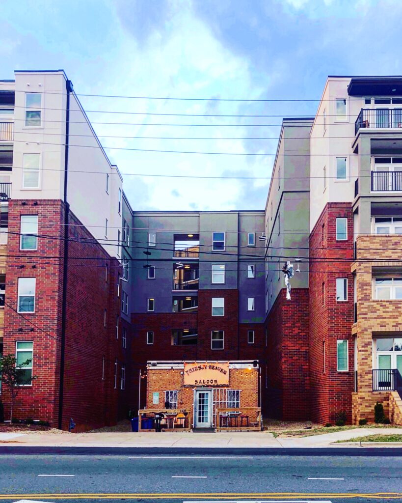 A small brick saloon with a wooden sign reading Thirsty Beaver Saloon sits between two large, modern apartment buildings under a partly cloudy sky—one of the iconic things to do in Charlotte—with power lines crossing the scene.